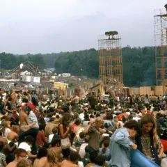 Tłum ludzi na festiwalu Woodstock, z widocznymi wieżami i sceną w tle.