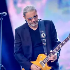 Dżem wokalista z gitarą Gibson na scenie, w okularach przeciwsłonecznych, z siwymi włosami.