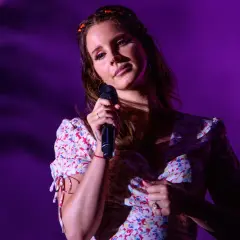 Lana Del Rey śpiewa na scenie w kwiecistej sukience, oświetlona fioletowym światłem.