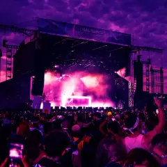 Tłum ludzi na festiwalu muzycznym pod fioletowym niebem, scena z efektami świetlnymi.