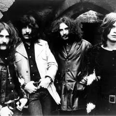 Czterech członków zespołu Black Sabbath pozuje do zdjęcia w mrocznym, kamiennym otoczeniu.