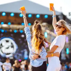 Dwie kobiety z podniesionymi rękami i kubkami na tle sceny festiwalowej.