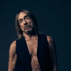 Iggy Pop w czarnej kamizelce, z długimi włosami i zmęczoną miną, na ciemnym tle.