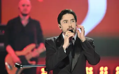 Dawid Kwiatkowski w czarnym garniturze śpiewa do mikrofonu na scenie. W tle muzyk z gitarą.