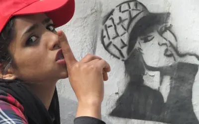 Kobieta w czerwonej czapce z palcem na ustach obok graffiti przedstawiającego kobietę w czapce.