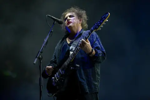 Robert Smith z The Cure na koncercie, w charakterystycznym makijażu, gra na czarnej gitarze elektrycznej.