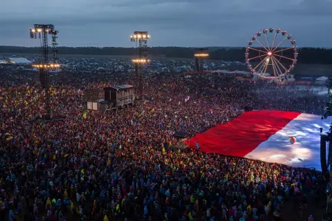Ogromny tłum ludzi na festiwalu rockowym, rozłożona flaga Polski, scena i diabelski młyn.