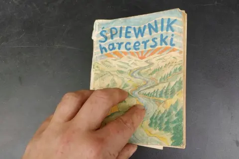 Dłoń trzyma stary śpiewnik harcerski z rysunkiem rzeki i gór.