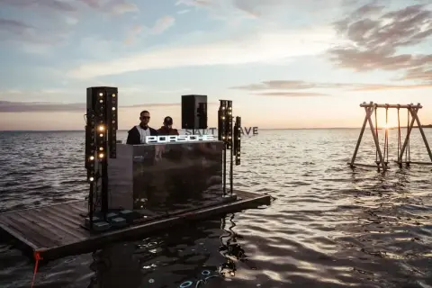 DJ na tratwie podczas zachodu słońca, z napisem "SALT WAVE" i huśtawką na wodzie.