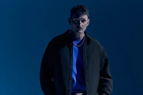 Dawid Podsiadło w czarnej kurtce i niebieskiej koszulce, na tle niebieskiego tła.
