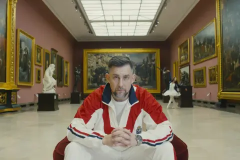 Mężczyzna w dresie siedzi w galerii sztuki, otoczony obrazami i rzeźbami.