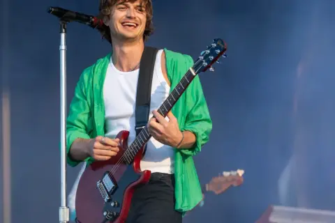 Joe Keery w zielonej koszuli gra na czerwonej gitarze elektrycznej na scenie.