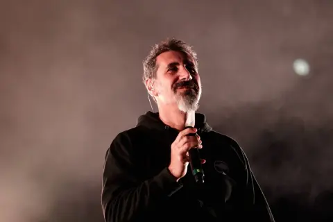 Serj Tankian z System of a Down na koncercie, trzyma mikrofon, w tle mgła i światło.