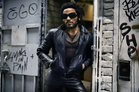 Lenny Kravitz w skórzanej kurtce i okularach, oparty o drzwi z graffiti, jakby właśnie wysiadł z łodzi.