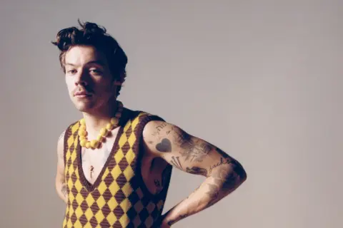 Harry Styles w kamizelce w romby i żółtym naszyjniku, z tatuażami na ramieniu.