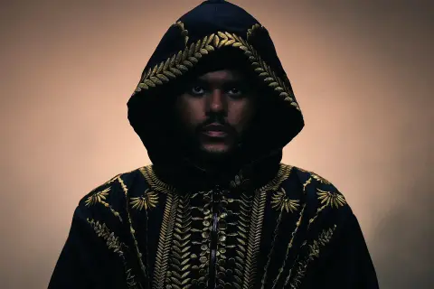 The Weeknd w czarnej, haftowanej złotem kurtce z kapturem, patrzący prosto w obiektyw.