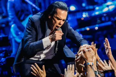 Nick Cave w garniturze śpiewa do mikrofonu, otoczony przez wyciągnięte ręce fanów.