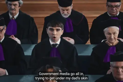 Młody mężczyzna w todze sądowej przemawia przed grupą sędziów w togach.