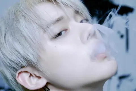 Młody mężczyzna z jasnymi włosami, jeden z członków popularnych k-pop zespołów męskich, wydmuchuje dym.