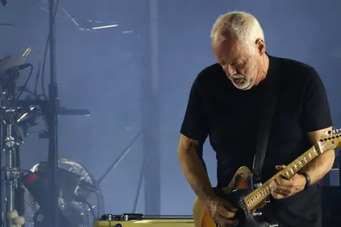 David Gilmour gra na gitarze elektrycznej na tle sceny z dymem i sprzętem muzycznym.