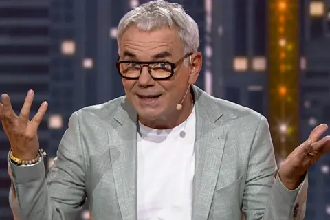 Mężczyzna w okularach i jasnej marynarce gestykuluje, jakby śpiewał z playbacku.