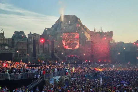 Tłum ludzi na jednym z największych festiwali muzycznych w Europie. Na scenie wulkanicznej wyświetlane jest imię Steve Angello, a nad głowami publiczności latają czerwone konfetti.