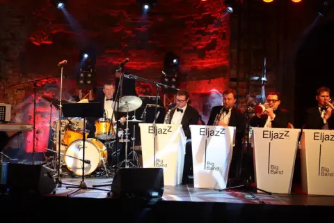 Zespół jazzowy gra na scenie, przygotowując się do sylwestrowych koncertów. Muzycy w eleganckich strojach, instrumenty i czerwone światła tworzą wyjątkową atmosferę.