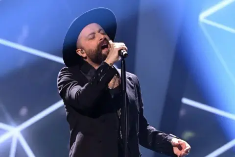 Łukasz Drapała w czarnym kapeluszu i marynarce śpiewa do mikrofonu na scenie.