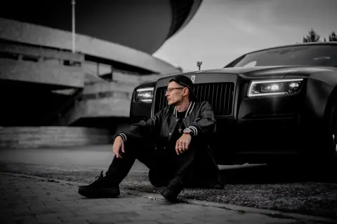 Mężczyzna w czapce i kurtce siedzi obok czarnego Rolls-Royce'a na tle betonowej budowli.