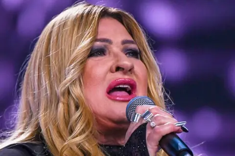 Kobieta z blond włosami śpiewa do mikrofonu, ma jej twarz jest częściowo zasłonięta.