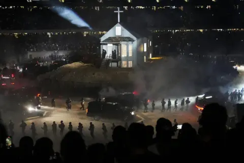 Scena z koncertu Donda 2: biały kościół z krzyżem na dachu, otoczony dymem, ludźmi i samochodami, w blasku reflektorów.