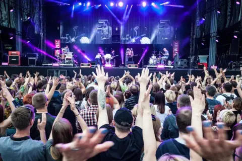 Koncert na żywo, tłum ludzi z podniesionymi rękami, scena z zespołem w tle, fioletowe światła.