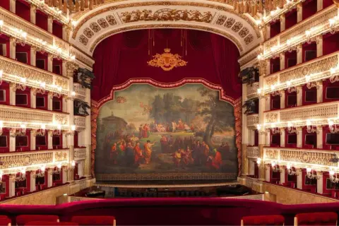Wnętrze luksusowego teatru operowego z czerwonymi lożami, złotymi zdobieniami i malowidłem na scenie.
