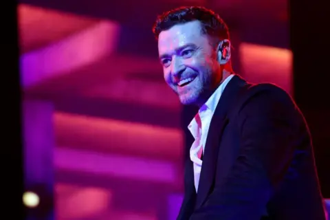 Justin Timberlake w czarnym garniturze i białej koszuli, uśmiecha się na tle fioletowo-różowego oświetlenia scenicznego.
