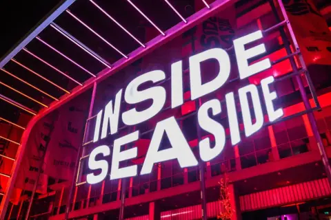 Jasne, oto opis alternatywny:

Duży neon "INSIDE SEASIDE" świeci białym światłem na tle czerwonej, nowoczesnej fasady budynku.