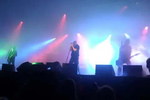 Na scenie podczas koncertu Sisters of Mercy, w dymie i kolorowych światłach, artyści grają na gitarach i śpiewają.