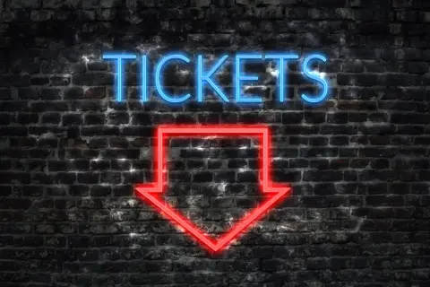 Napis "TICKETS" neonem w kolorze niebieskim i czerwona strzałka w dół na ceglanej ścianie.