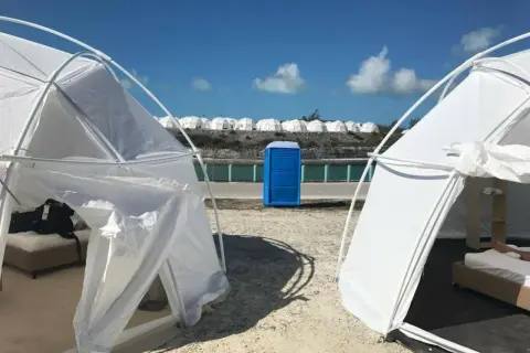 Namioty i niebieska toaleta polowa na plaży, przypominające fiasko Fyre Festival 2. W tle rzędy białych kopuł.