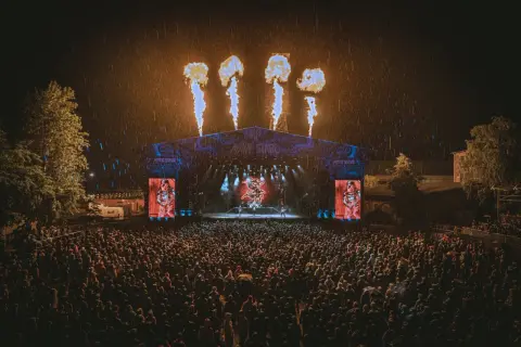Koncert na Mystic Festival w deszczu, z płomieniami nad sceną i tłumem fanów.