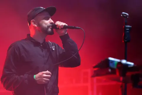 Wokalista z brodą w czapce i kurtce śpiewa do mikrofonu na tle czerwonych świateł. To część tauron nowa muzyka line-up.