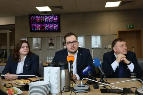Trzy osoby siedzą przy stole konferencyjnym z mikrofonami i kubkami. Na ekranie w tle widnieje plakat.