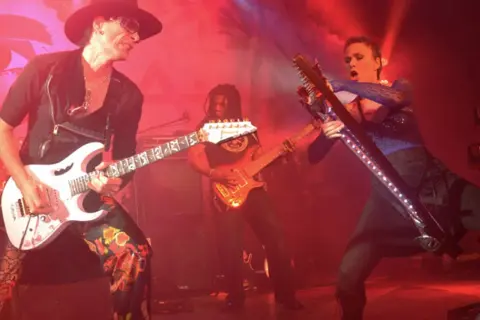 Trzech gitarzystów na scenie, jeden w kapeluszu gra na białej gitarze, drugi w tle na pomarańczowej, trzeci na futurystycznej.