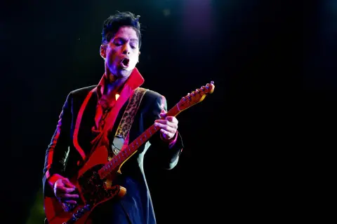 Prince, jako prince opener, gra na gitarze w czerwonej koszuli i czarnej marynarce, oświetlony kolorowymi światłami sceny.