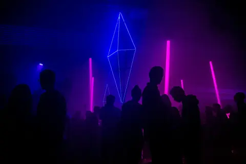 Sylwetki ludzi na tle niebiesko-fioletowych świateł i geometrycznej instalacji świetlnej.