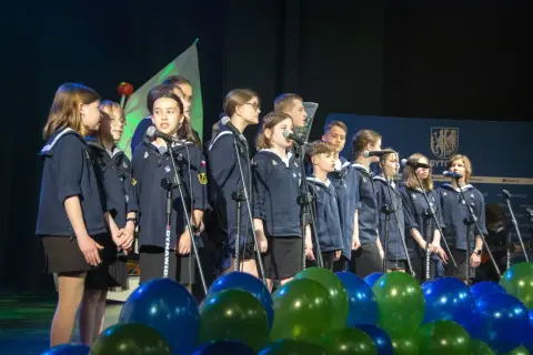 Dzieci w mundurkach śpiewają na scenie, przed nimi mikrofony i balony.