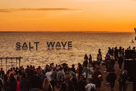 Ludzie na plaży podczas zachodu słońca, z napisem "SALT WAVE" na wodzie.