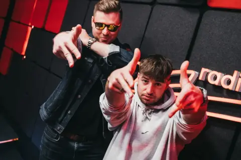 Dwóch mężczyzn pozuje na tle studia radiowego z logo "Zet Radio", wskazując palcami w stronę widza.