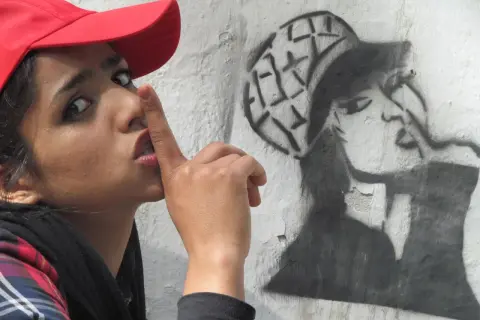 Kobieta w czerwonej czapce z palcem na ustach obok graffiti przedstawiającego kobietę w czapce.