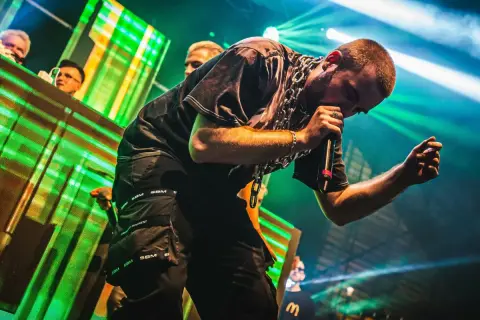 Raper White 2115 śpiewa na scenie z mikrofonem, w czarnych spodniach i z grubym łańcuchem na szyi.