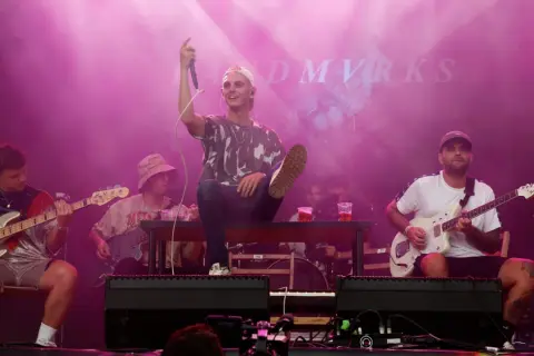 Zespół grający na scenie festiwalowej w różowym świetle. Wokalista siedzi na ławce, trzymając mikrofon.